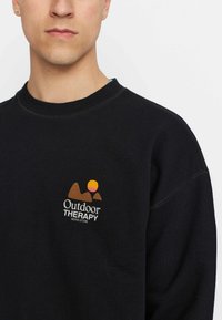 Zwarte sweatshirt met geribbelde ronde hals. Heeft een geborduurd logo in oranje, bruin en crème dat bergen en een zon afbeeldt.