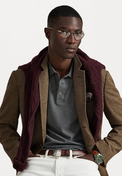 Καφέ σακάκι σε σχέδιο χερροβόρντ επικαλυμμένο από γκρι πόλο, με ένα μπορντό πλεκτό πουλόβερ ριγμένο στους ώμους. Λευκά παντελόνια, καφέ ζώνη και πράσινο ρολόι.