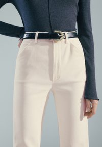 Personne portant un pantalon en velours côtelé crème taille haute avec une ceinture en cuir noire et un haut ajusté à manches longues foncé, posant devant un fond uni.