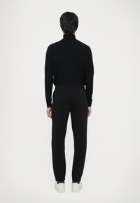 Pessoa de costas, vestindo uma blusa preta de mangas longas, calças pretas e ténis brancos, num fundo claro e liso.