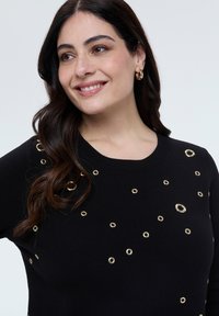 Lächelnde Frau mit langen dunklen Haaren trägt schwarzen Pullover, der mit verstreuten goldenen Ösen verziert ist, und kleine goldene Creolen vor weißem Hintergrund.