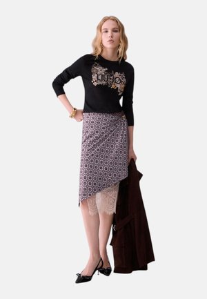 Donna che indossa un top nero a maniche lunghe con logo floreale, gonna a portafoglio fantasia con dettaglio in pizzo, tiene in mano un cappotto scuro e tacchi neri a punta.