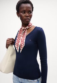 Top din tricot cu maneci lungi de culoare bleumarin, cu decolteu în V, asortat cu o eșarfă crem cu modele florale roșii. Modelul ține o geantă albă.