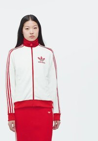 Bijela i crvena Adidas trenirka s visokim ovratnikom, koja ima tri crvene pruge na rukavima i logo na prsima, u kombinaciji s crvenom suknjom.