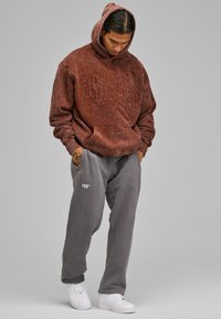 Felpa oversize marrone con testo in rilievo, abbinata a pantaloni della tuta grigi con un piccolo logo e sneakers bianche. Tessuto morbido.