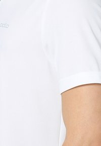 Gros plan d'une personne portant un maillot de sport blanc texturé à manches courtes avec un petit logo sur la poitrine.