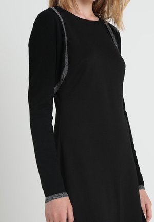 Femme portant une robe noire à manches longues avec un bord argenté sur les épaules et les poignets, debout devant un fond clair uni.