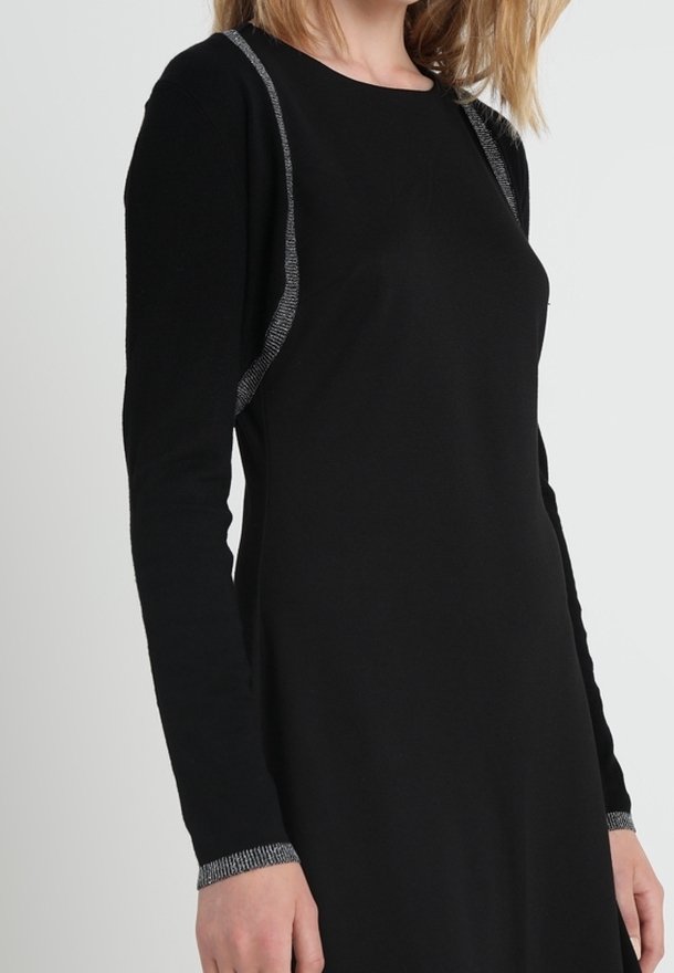 Femme portant une robe noire à manches longues avec un bord argenté sur les épaules et les poignets, debout devant un fond clair uni.