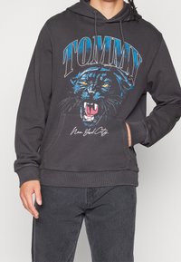 Tumma harmaa huppari, joka on valmistettu puuvillasekoitteesta. Siinä on sininen ja keltainen pantterigrafiikka, etutasku sekä ribbatut hihansuut ja helma.