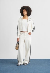 Fehér sportdzseki fekete csíkokkal, hozzáillő fehér felső, bőszárú fehér nadrág és ezüst cipő. Kis fekete arany táska.