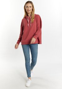 Felpa color ruggine con scollatura a cordoncino, vestibilità ampia, maniche lunghe, abbinata a jeans skinny blu e sneakers bianche. Sfondo semplice.