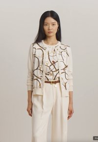Cardigan alb cu un model din lanțuri în nuanțe de aur și maro, asociat cu un top alb asortat și pantaloni largi de crem, accentuați de o curea maro.