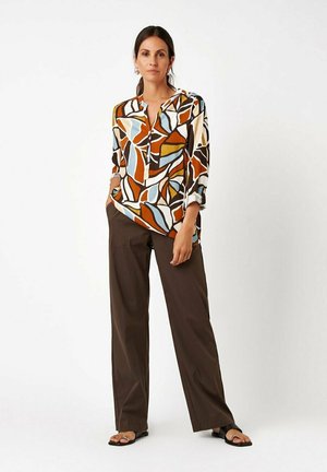 Woman standing with hands in pockets wearing a multicolored abstract print blouse, brown wide-leg pants, and black sandals on a white background.