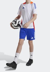 Valkoinen urheilupaita oransseilla ja sinisillä tehosteilla, siniset shortsit ja mustat nappikset. Henkilöllä on kädessään mustavalkoinen jalkapallo.