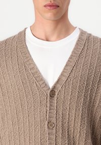 Beige kötött kardigán V-nyakkal, texturált mintával, bordázott szegéllyel és két barna gombbal az elején.