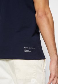 Gros plan d'une personne portant une chemise bleu marine avec le texte blanc "French Sportswear est. nineteen twenty seven" et un pantalon écru.