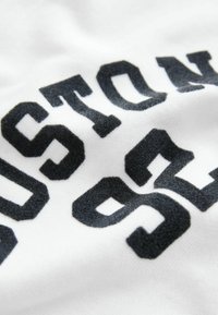 Detailaufnahme von schwarzen Varsity-Buchstaben und -Zahlen "STAN" und "951" auf weißem Stoff, die Textur und Nähdetails zeigen.