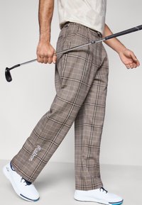 Καφέ καρό παντελόνια γκολφ με εφαρμοστή ραφή, με διακριτικό κεντημένο λογότυπο στο κάτω μέρος του ποδιού, σε συνδυασμό με λευκά αθλητικά παπούτσια.