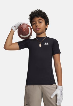 Mustas Under Armour särgis ja beežides lühikestes pükstes poiss hoiab jalgpalli õlal, kannab valgeid kindaid ja ripatsiga kaelakeed.