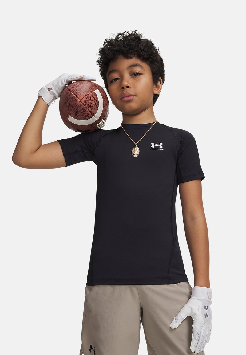 Junge in schwarzem Under Armour-Shirt und beigen Shorts hält einen Football auf der Schulter, trägt weiße Handschuhe und eine Halskette mit Anhänger.