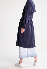 Tmavě modrý dlouhý kabát s uvázaným pasem, širokými rukávy a vrstveným zadním designem, který je skombinován se světle modrými zkrácenými širokými kalhotami.