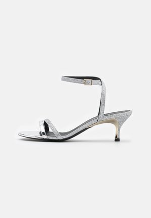 Sandales à talons en paillettes argentées avec deux fines brides et une bride de cheville dotée d'une boucle dorée. Talon stiletto avec une semelle noire.