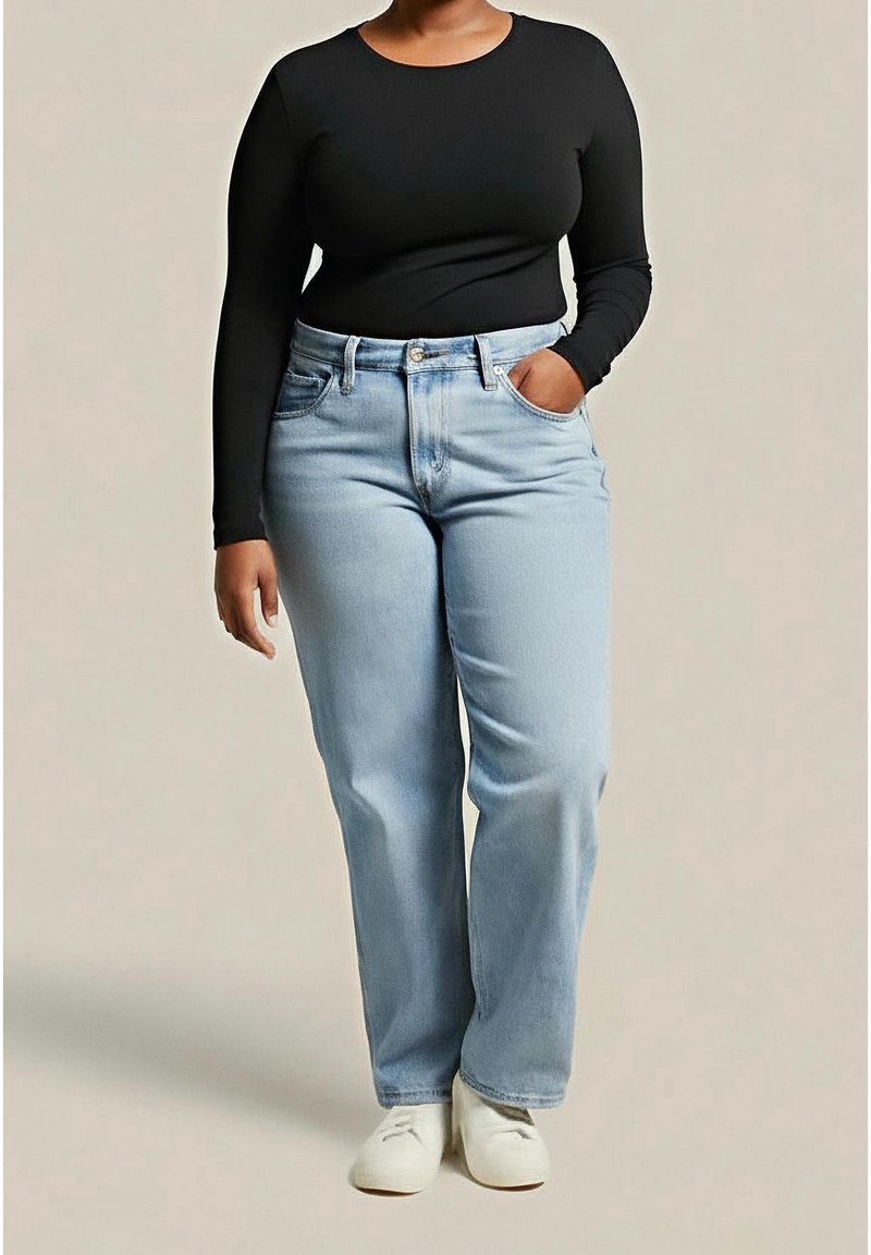 Femme portant une chemise noire à manches longues, un jean bleu clair droit et des chaussures blanches, une main dans la poche, debout sur un fond beige.