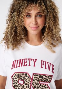 Une femme aux cheveux blonds bouclés sourit, portant une chemise blanche avec "NINETY FIVE" et le numéro 95 en imprimé léopard sur le devant.