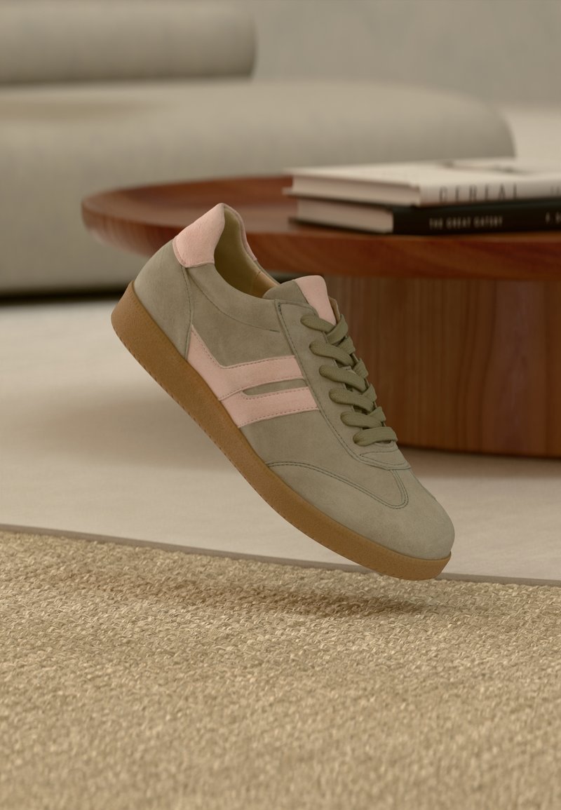 Baskets grises décontractées avec des accents roses et une semelle en gomme flottant au-dessus d'un tapis beige tissé, d'une table basse en bois et de livres empilés dans un salon aux tons neutres.