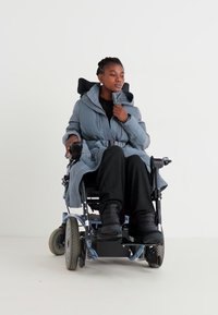 Moteris pilko pūkuoto paltuko su diržu sėdi elektriniame vežimėlyje, vilkėdama juodas kelnes ir batus prieš baltą foną.