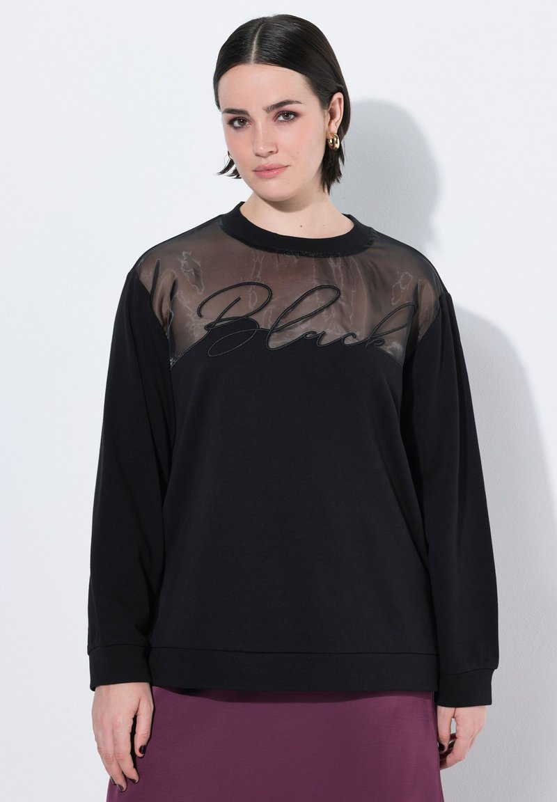 Svart sweatshirt med ett genomskinligt nätpanel högst upp, med ordet "Black" i kursiv. Långa ärmar och avslappnad passform.