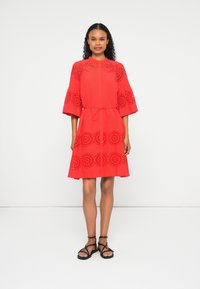 Mulher vestindo um vestido vermelho até os joelhos com padrões recortados circulares, mangas 3/4, um cinto na cintura e sandálias planas pretas com tiras, de pé contra um fundo branco.