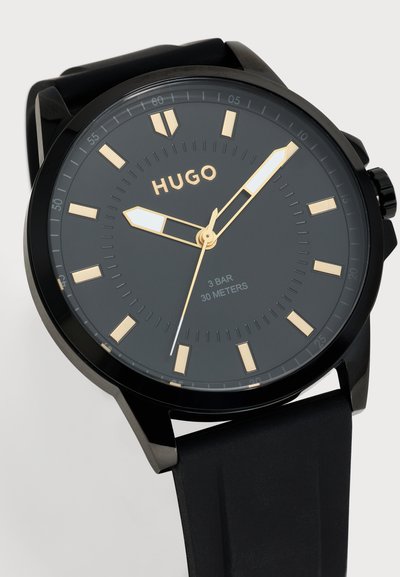 Montre-bracelet Hugo noire avec un cadran gris foncé, des aiguilles et des index de couleur dorée, et un bracelet noir ; indiquée 3 BAR, 30 mètres.