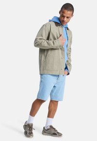 Ανοιχτό πράσινο μπουφάν με γιακά, δύο τσέπες στο στήθος, φορεμένο πάνω από ένα ανοιχτό μπλε φούτερ με κουκούλα. Συνοδεύεται από ανοιχτό μπλε σορτς και γκρι αθλητικά παπούτσια.