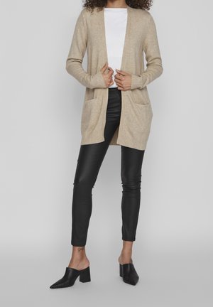 Beige cardigan met lange mouwen, open voorkant, twee zakken en ribbelachtige zoom. Gecombineerd met zwarte leren broek en zwarte muiltjes.
