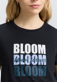 T-shirt noir avec un imprimé graphique affichant le mot "BLOOM" dans des tailles et des nuances de blanc et de bleu variées, avec "bloom forever" en police cursive.