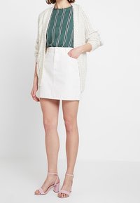 Krämfärgad tjockstickad cardigan, grönrandig blus, vit denim minikjol, ljusrosa klacksandaler. Modern, avslappnad outfitdesign.