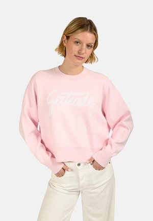 Femme aux cheveux blonds courts portant un sweat-shirt rose clair et un pantalon blanc, debout les mains dans les poches devant un fond uni.