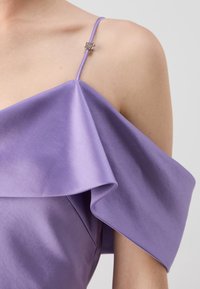 Zbliżenie na lawendową sukienkę bez ramiączek z cienkim paskiem i dekoracyjną srebrną koraliką na ramieniu na neutralnym tle.