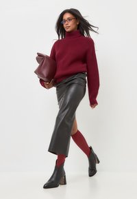 Bordeauxfarbener Strick-Turtleneck-Pullover, schwarzer Lederrock mit Seitenschlitz, rote Strumpfhosen und schwarze Stiefeletten mit gemustertem Absatz, dazu eine passende Tasche.