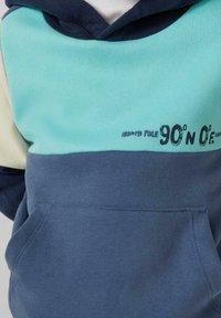 Mikina s farebnými blokmi v svetlo modrej a námorníckej farbe, predné vrecko a nápis "Severný pól 90°S 0°V" potlačený na hrudi.
