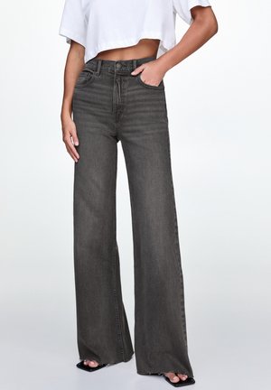 Femme portant un jean noir délavé taille haute, jambes larges, associé à un t-shirt blanc court, debout avec une main dans la poche.