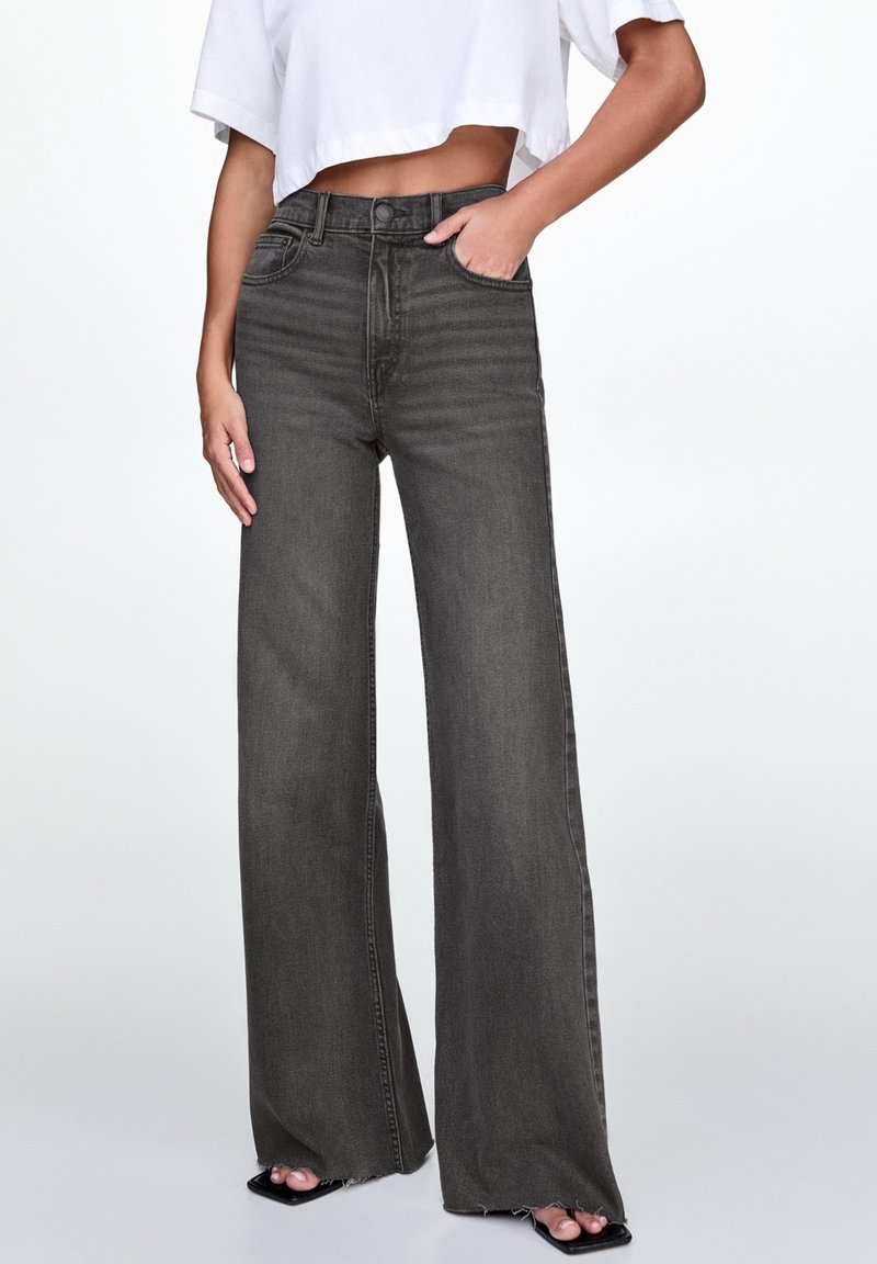 Femme portant un jean noir délavé taille haute, jambes larges, associé à un t-shirt blanc court, debout avec une main dans la poche.