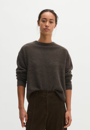 Femme portant un pull à manches longues marron foncé et un pantalon taille haute marron foncé, debout sur un fond blanc uni.