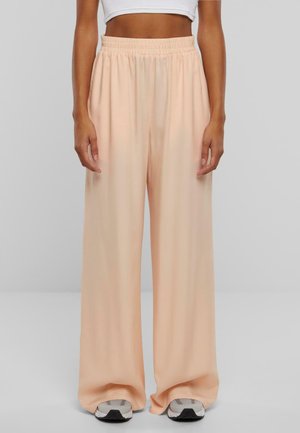 Mujer con pantalones anchos y sueltos de color melocotón con cintura elástica y zapatillas blancas, camiseta blanca corta visible por encima de la cintura.