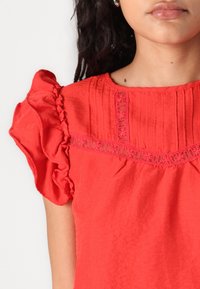 Rode blouse met opgeblazen mouwen, gestructureerde stof en decoratieve kanten rand langs de halslijn en het lijfje. Lichte rimpels zichtbaar.