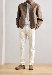 Brun lätt jacka med elastiska ärmar, grå t-shirt, vita raka byxor och bruna mocka slip-on skor på ett trägolv.