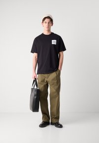 Zwarte katoenen T-shirt met logo patch, olijfgroene broek, zwarte leren schoenen en een zwarte tote bag. Model staat tegen een effen achtergrond.