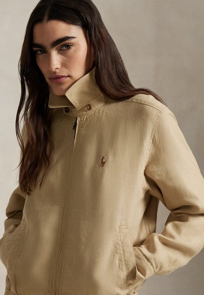 Beige, lettvekt jakke med høy krage og glidelås. Har sidelommer og en liten logo på brystet. Glatt tekstur.