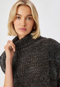 Dunkler, strukturierter Strickpullover mit hohem Kragen und offenem Webmuster, versehen mit metallischen Akzenten. Die Hand des Models befindet sich in der Nähe des Kragens.